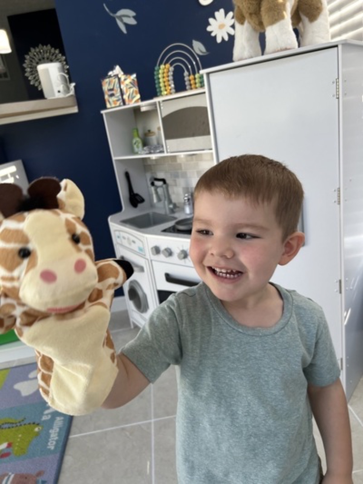 Child smiling with a giraffe puppet in the play kitchen area
